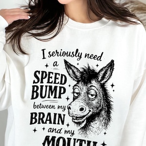 Peut inclure: Sweat-shirt blanc avec l'inscription noire "I seriously need a SPEED BUMP between my BRAIN and my MOUTH". Une illustration en noir et blanc d'un âne souriant est à droite. Le sweat-shirt est de style décontracté.