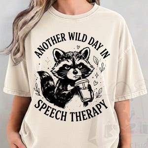 Peut inclure: Un t-shirt à manches courtes de couleur crème avec un motif noir représentant un raton laveur buvant dans un gobelet avec une paille. Le texte "ANOTHER WILD DAY IN SPEECH THERAPY" est imprimé en lettres grasses et arquées.