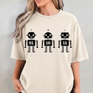 Peut inclure: T-shirt crème avec trois motifs de robots noirs. Chaque robot affiche un nombre binaire différent : 01, 10 et 11. Les robots ont un design simple et géométrique avec une tête, un corps et des jambes. Le t-shirt est de style décontracté et unisexe.