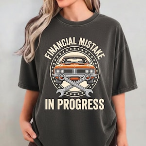 Peut inclure: T-shirt gris foncé avec un motif graphique représentant une voiture classique orange, des clés croisées et le texte "FINANCIAL MISTAKE IN PROGRESS" dans un cercle. Le t-shirt a des manches courtes et un col rond.