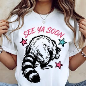 Puede incluir: Camiseta blanca con una ilustración de mapache en blanco y negro, estrellas rosas y turquesas, y el texto rosa "SEE YA SOON". El mapache tiene una cola a rayas y está de espaldas. Una camiseta gráfica informal.