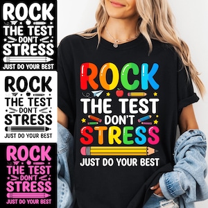 May include: Black t-shirt featuring the phrase "ROCK THE TEST DON'T STRESS JUST DO YOUR BEST" in vibrant colours. The design incorporates a pencil, apple, and paper aeroplane. A casual wear item.