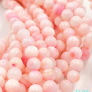 May include: Close-up of a strand of pink gemstone beads. The beads are round and have a soft, pearly luster. The color varies from pale pink to deeper rose, creating a subtle gradient. The beads are strung together, suggesting they are for jewelry making.