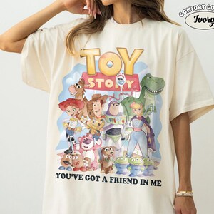 May include: Ivory-colored t-shirt featuring a colorful graphic of Toy Story characters. The design includes Woody, Buzz Lightyear, and other characters from the movie, with the text "TOY STORY" and "YOU'VE GOT A FRIEND IN ME".