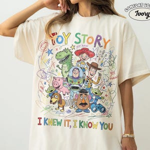 Puede incluir: Una camiseta de color marfil con un gr&aacute;fico colorido de los personajes de Toy Story, incluyendo a Woody, Buzz Lightyear, Rex y el Sr. Patata. El dise&ntilde;o incluye l&iacute;neas estilo garabato, estrellas y el texto "TOY STORY" e "I KNEW IT, I KNOW YOU."