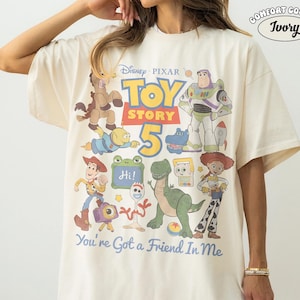 May include: Ivory-colored t-shirt featuring a graphic of characters from the Disney Pixar movie Toy Story 5. The design includes Buzz Lightyear, Woody, and other characters, with the text "You're Got a Friend In Me."