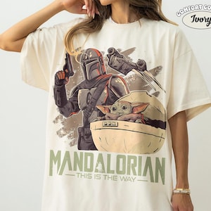 May include: An ivory t-shirt featuring a graphic print of a Mandalorian character holding a blaster, a small creature in a floating pod, and a starship. The text reads "MANDALORIAN" and "THIS IS THE WAY" in green, with a "COMFORT COLORS IVORY" logo.