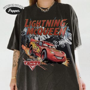 May include: A charcoal gray short-sleeve t-shirt featuring a graphic of the character Lightning McQueen from the Disney Pixar movie Cars. The design includes the text Lightning McQueen and the Cars logo, with a distressed, faded aesthetic.