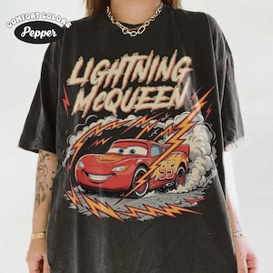 May include: Dark gray t-shirt featuring Lightning McQueen from the Cars movie. The design includes the text "LIGHTNING McQUEEN" above a red race car with the number 95. The shirt has a vintage, distressed look.