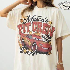 May include: Ivory-colored t-shirt with the text "Mason's Pit Crew" in red and yellow. A red race car with the number 95 is featured on the front, along with flames and a checkered flag design. The shirt is a Comfort Colors brand.