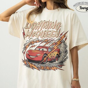 May include: Ivory-colored t-shirt featuring a graphic of Lightning McQueen from the Cars movie. The design includes the text "Lightning McQueen" above the car, with lightning bolts and smoke effects. The number "95" is visible on the car.