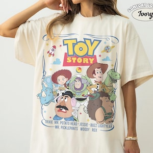 May include: Ivory-colored t-shirt featuring a colorful graphic of Toy Story characters. The design includes the words "TOY STORY" in a playful font, along with character names like Woody, Buzz Lightyear, and Jessie. The shirt is labeled "COMFORT COLORS" and "Ivory".