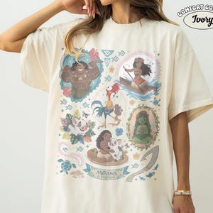 May include: Ivory Comfort Colors t-shirt featuring a colorful graphic of characters from the animated film Moana. The design includes illustrations of Moana, Maui, Pua, Hei Hei, and other elements from the movie. The text "MOANA & FRIENDS" is also included.