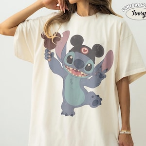 May include: An ivory t-shirt featuring a graphic of the blue character Stitch wearing black mouse ears and holding a brown ice cream bar. The text COMFORT COLORS IVORY is printed in the top right corner. The shirt has short sleeves and a crew neck.