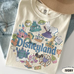 May include: Cream-coloured Comfort Colors t-shirt with a Disneyland graphic. The design features the castle, Matterhorn, and a Ferris wheel, with the word "Disneyland" in blue script. The shirt is ivory in colour.
