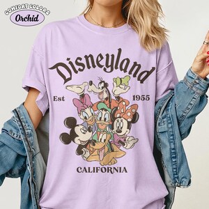 May include: A light purple t-shirt with the word "Disneyland" in brown script, featuring cartoon characters. The shirt also includes the text "Est 1955 CALIFORNIA" in brown. The shirt is made of Comfort Colors Orchid material.