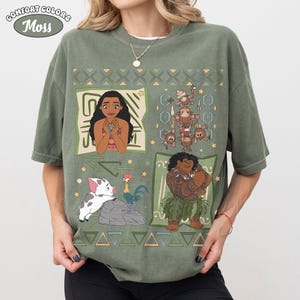 May include: A moss green short-sleeve t-shirt featuring a graphic print with four distinct panels. The design includes Moana, Maui, Pua the pig, Heihei the rooster, and a group of Kakamora characters, all surrounded by geometric patterns.