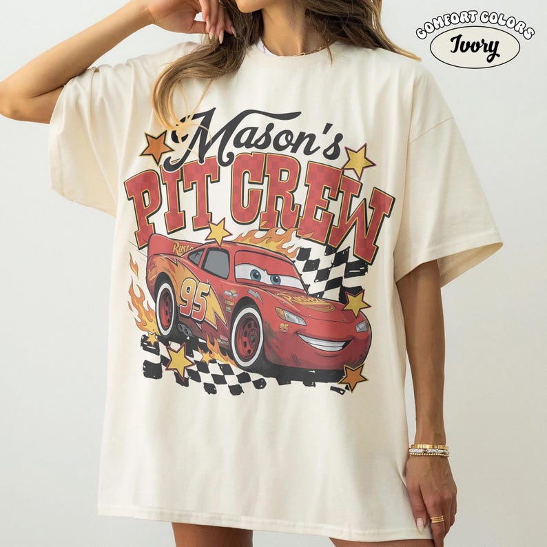 May include: Ivory-colored t-shirt with the text "Mason's Pit Crew" in red and yellow. A red race car with the number 95 is featured on the front, along with flames and a checkered flag design. The shirt is a Comfort Colors brand.