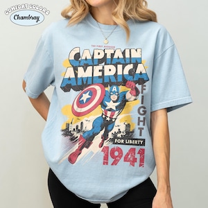May include: A light blue short-sleeve t-shirt featuring a graphic of Captain America with a shield, city skyline, and the text "The First Avenger Captain America Fight For Liberty 1941". The top left corner includes a "Comfort Colors Chambray" logo.