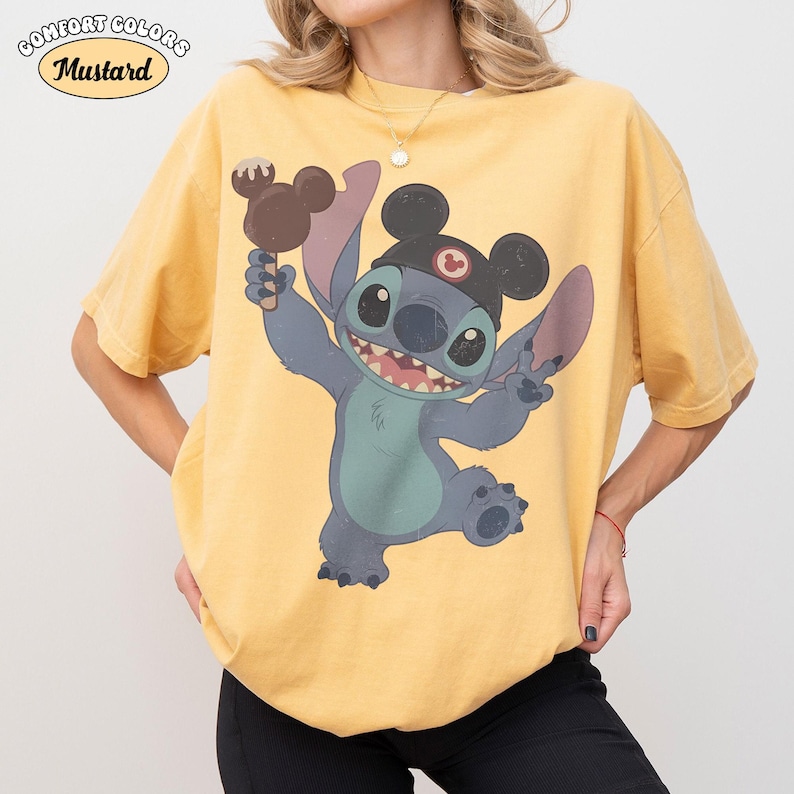 May include: A mustard yellow short-sleeve t-shirt featuring a graphic of the character Stitch wearing black mouse ears and holding a brown chocolate-covered treat. The text Comfort Colors and Mustard is printed in the upper left corner of the image.