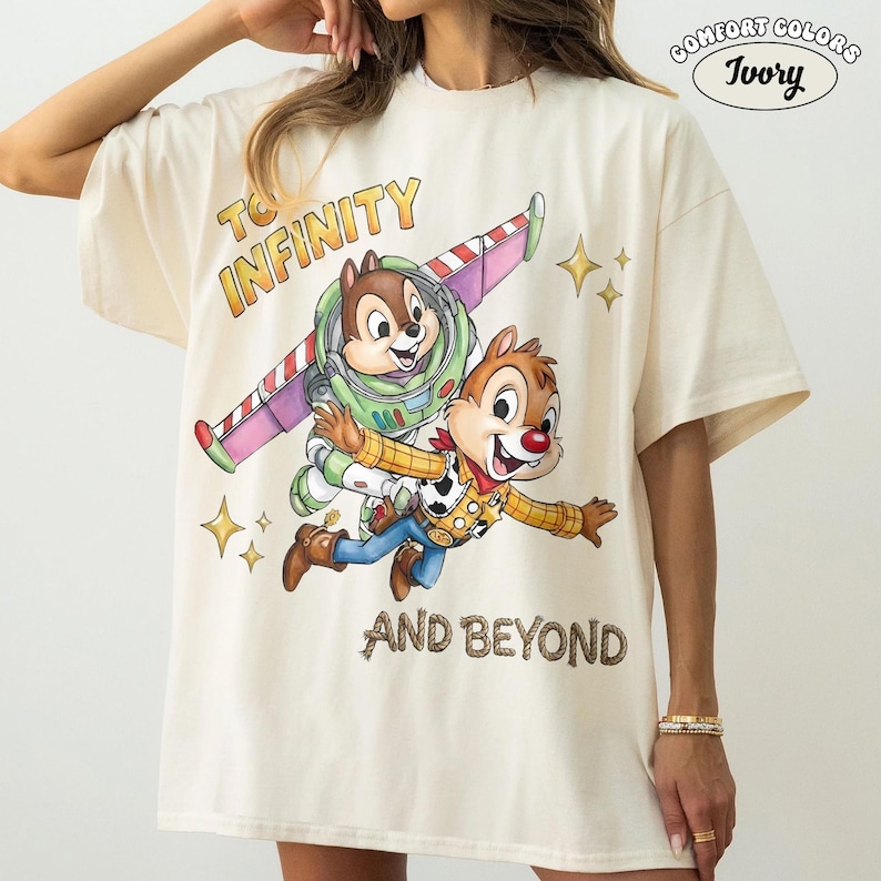 May include: Ivory-colored t-shirt featuring a graphic of Chip and Dale as Buzz Lightyear and Woody, with the text "TO INFINITY AND BEYOND." The shirt has a relaxed fit and is made of Comfort Colors material.