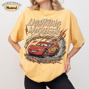 May include: Mustard-colored t-shirt featuring a graphic of Lightning McQueen from the Cars movie. The design includes the text "LIGHTNING McQUEEN" and the number "95" on the red race car, with lightning bolts and smoke effects.