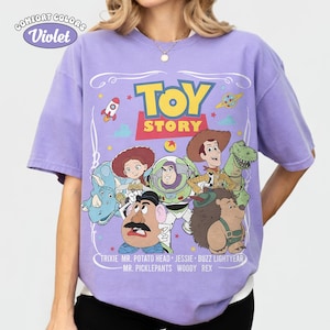 May include: A violet Comfort Colors t-shirt featuring a graphic of Toy Story characters. The design includes the words "TOY STORY" in yellow and red, with character names below. The shirt has short sleeves.
