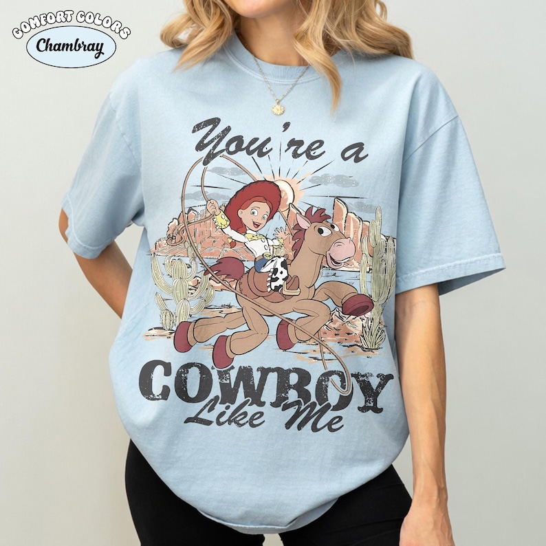 May include: A light blue short-sleeve t-shirt featuring a graphic of a cowgirl riding a horse in a desert landscape with cacti and mountains. The text reads "You're a COWBOY Like Me" and "COMFORT COLORS Chambray" is printed in the upper left corner.