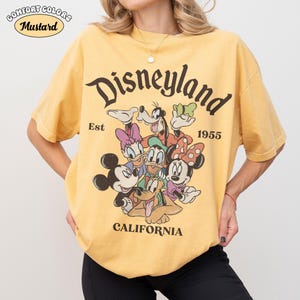 May include: Mustard yellow t-shirt with a Disneyland graphic featuring Mickey Mouse, Minnie Mouse, Donald Duck, Daisy Duck, Goofy, and Pluto. The text "Disneyland California Est 1955" is printed on the front. The shirt is a Comfort Colors brand.