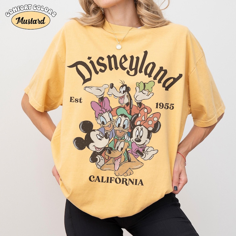 May include: Mustard yellow t-shirt with a Disneyland graphic featuring Mickey Mouse, Minnie Mouse, Donald Duck, Daisy Duck, Goofy, and Pluto. The text "Disneyland California Est 1955" is printed on the front. The shirt is a Comfort Colors brand.