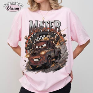 May include: A light pink Comfort Colors t-shirt featuring a graphic of Mater from the movie Cars. The graphic includes the word "MATER" in a distressed font, a tire, and lightning bolts. The shirt has short sleeves.