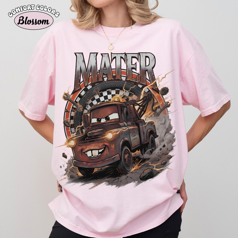 May include: A light pink Comfort Colors t-shirt featuring a graphic of Mater from the movie Cars. The graphic includes the word "MATER" in a distressed font, a tire, and lightning bolts. The shirt has short sleeves.