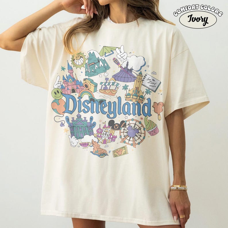 May include: Ivory-coloured t-shirt featuring a Disneyland graphic. The design includes park icons like a castle, teacup ride, and the Disneyland logo. The shirt is labelled "Comfort Colors" and "Ivory".