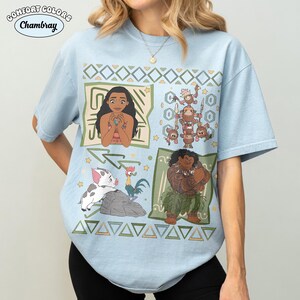 May include: A light blue short-sleeve t-shirt featuring a graphic print of characters from the movie Moana, including Moana, Maui, Pua, Heihei, and the Kakamora. The design includes geometric patterns and small stars on a light blue fabric.