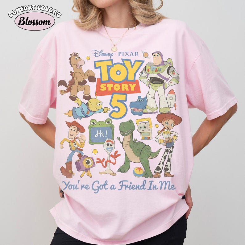 May include: Pink Comfort Colors t-shirt featuring a colorful graphic of Toy Story characters and the text "Toy Story 5". The design includes Woody, Buzz Lightyear, and other characters from the animated film. The text "You're Got a Friend In Me" is also included.