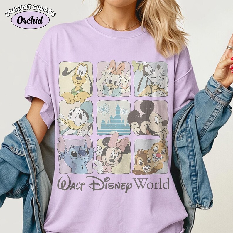 Op de afbeelding: Een lichtpaars t-shirt met een raster van negen vierkanten met diverse Disney-figuren en een kasteel, waaronder Pluto, Katrien Duck, Goofy, Donald Duck, Mickey Mouse, Stitch, Minnie Mouse en Knabbel en Babbel, met de tekst Walt Disney World.