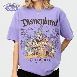 May include: A violet Comfort Colors t-shirt featuring the word "Disneyland" in brown, with a graphic of Disney characters and the text "CALIFORNIA EST. 1955". The shirt has short sleeves and a crew neck.