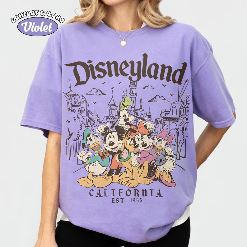 May include: A violet Comfort Colors t-shirt featuring the word "Disneyland" in brown, with a graphic of Disney characters and the text "CALIFORNIA EST. 1955". The shirt has short sleeves and a crew neck.