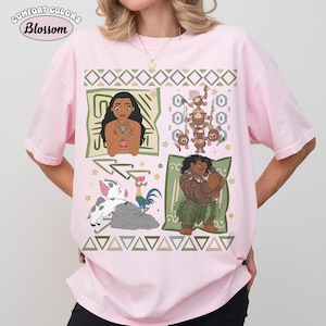 May include: A light pink short-sleeve t-shirt featuring a graphic print of characters from the movie Moana, including Moana, Maui, Pua, Heihei, and the Kakamora. The design includes geometric patterns, stars, and the text Comfort Colors Blossom.