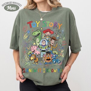 Puede incluir: Una camiseta de manga corta color verde musgo con un gr&aacute;fico colorido de los personajes de Toy Story, incluyendo a Woody, Buzz Lightyear, Jessie, Rex, Hamm, Slinky Dog, Mr. Potato Head y alien&iacute;genas. El dise&ntilde;o incluye garabatos y el texto TOY STORY.