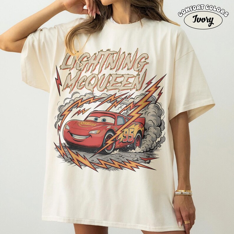 May include: Ivory-colored t-shirt featuring a graphic of Lightning McQueen from the Cars movie. The design includes the text "Lightning McQueen" above the car, with lightning bolts and smoke effects. The number "95" is visible on the car.