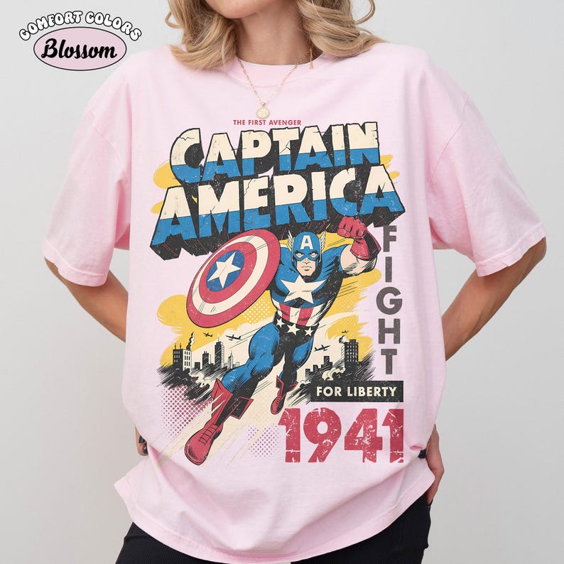 May include: A light pink short-sleeve t-shirt featuring a graphic of Captain America with a shield, the text THE FIRST AVENGER CAPTAIN AMERICA FIGHT FOR LIBERTY 1941, and a city skyline background in blue, red, white, and yellow colors.