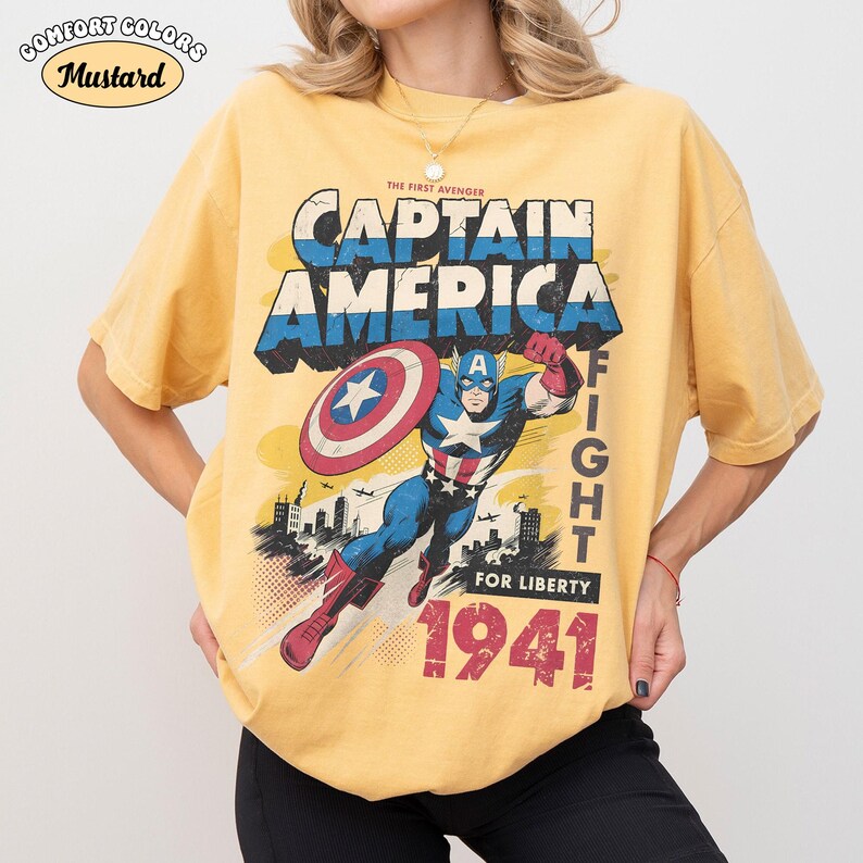 May include: A mustard yellow short-sleeve t-shirt featuring a graphic print of Captain America in a dynamic pose with a shield. The text reads "THE FIRST AVENGER CAPTAIN AMERICA FIGHT FOR LIBERTY 1941". A small logo in the top left corner says "COMFORT COLORS MUSTARD".