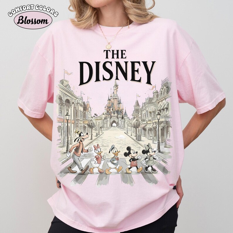May include: Pink t-shirt with the text "THE DISNEY" above a graphic of Disney characters crossing a street in front of a castle. The shirt has the "Comfort Colors" and "Blossom" logos.