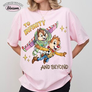 May include: Pink Comfort Colors t-shirt featuring Chip and Dale as Buzz Lightyear and Woody, with the text "TO INFINITY AND BEYOND". The design includes stars and a colorful spaceship wing. The shirt has a Blossom logo in the upper left corner.