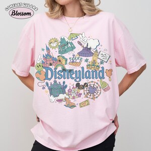 May include: Pale pink Comfort Colors t-shirt featuring a Disneyland graphic. The design includes Disneyland icons and the word "Disneyland" in blue. The shirt has short sleeves and is made of a soft material.