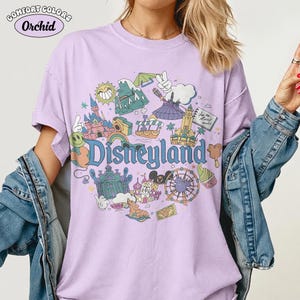 May include: A lilac t-shirt with a Disneyland graphic. The graphic features various park icons in a circular design, including a castle, a Ferris wheel, and the word "Disneyland". The shirt has the words "Comfort Colors" and "Orchid" in the upper left corner.