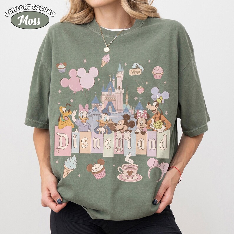 May include: Moss green Comfort Colors t-shirt featuring a Disneyland graphic. The design includes cartoon characters, a castle, and the word "Disneyland" in pastel colors. Additional elements include ice cream cones, cupcakes, and Mickey Mouse ears.