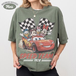May include: Moss green Comfort Colors t-shirt with a graphic of Disney characters in a red race car with checkered flags. The car has the number 95 on it, and the text "SPEED TEAM 1928" is below the car.