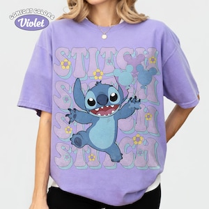 May include: A violet short-sleeve t-shirt featuring a graphic of the blue character Stitch holding light blue balloons. The background includes repeating text that reads STITCH in a wavy font, accented with small yellow and light blue flowers.
